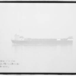 Schwarzweißaufnahme eines Frachtschiffs in dichtem Nebel über ruhigem Wasser mit schwacher Spiegelung, sichtbarem Wasserzeichen des Internationalen Maritimen Museums Hamburg unten links und Filmrand um das Bild.