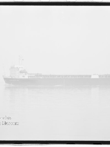 Schwarzweißaufnahme eines Frachtschiffs in dichtem Nebel über ruhigem Wasser mit schwacher Spiegelung, sichtbarem Wasserzeichen des Internationalen Maritimen Museums Hamburg unten links und Filmrand um das Bild.