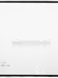 In dichtem Nebel zeichnet sich ein schwach sichtbares Frachtschiff auf ruhigem Wasser mit dezenter Spiegelung ab; das hochbelichtete Schwarzweißbild zeigt einen schwarzen Filmrand und ein Wasserzeichen unten links.