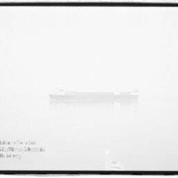 In dichtem Nebel zeichnet sich ein schwach sichtbares Frachtschiff auf ruhigem Wasser mit dezenter Spiegelung ab; das hochbelichtete Schwarzweißbild zeigt einen schwarzen Filmrand und ein Wasserzeichen unten links.