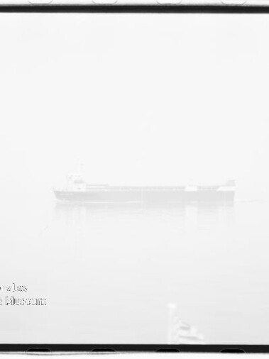 In dichtem Nebel zeichnet sich ein schwach sichtbares Frachtschiff auf ruhigem Wasser mit dezenter Spiegelung ab; das hochbelichtete Schwarzweißbild zeigt einen schwarzen Filmrand und ein Wasserzeichen unten links.