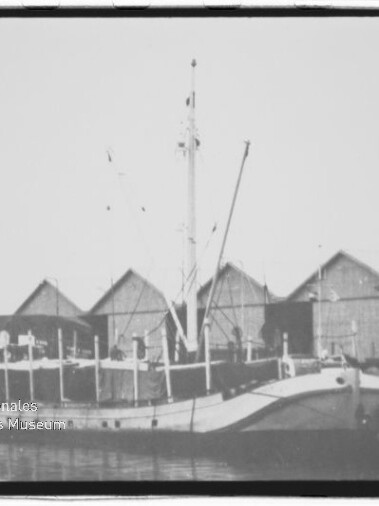 Schwarzweiß-Foto eines angelegten Frachtschiffs am Kai vor einer Reihe giebeldachiger Lagerhäuser und einem Haus, mit ruhiger Wasseroberfläche im Vordergrund.