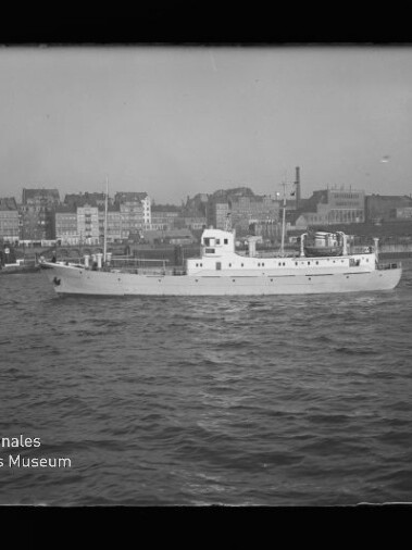 Weißes Motorpassagierschiff mit mittigem Aufbau fährt auf leicht bewegter Wasseroberfläche, im Hintergrund mehrstöckige Gebäude und Hafenbauten einer städtischen Uferzeile.