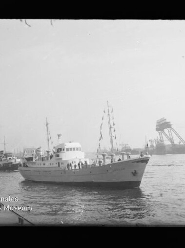 Schwarzweiß-Foto eines weißen Passagierboots mit Personen an Deck und Festtagswimpeln, das in einem Industriehafen zwischen weiteren Schiffen und großen Krananlagen fährt; unten links sichtbares Wasserzeichen des Internationalen Maritimen Museums Hamburg.