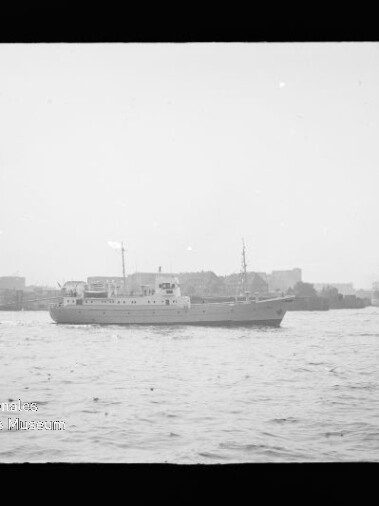 Schwarzweißaufnahme eines mittleren Schiffes, das in einem Hafenbecken vor einer städtisch-industriellen Kulisse mit weiteren Schiffen fährt; unten links sichtbar das Logo und der Schriftzug des Internationalen Maritimen Museums Hamburg.
