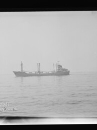 Schwarzweißaufnahme eines Frachtschiffs auf ruhiger See, im Vordergrund links zwei Männer auf einem kleinen Boot, mit schwarzem Bildrand und Wasserzeichen des Internationalen Maritimen Museums Hamburg unten links.