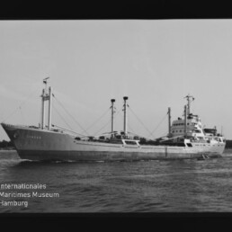 Schwarzweißaufnahme eines großen Frachtschiffs mit mehreren Masten und Ladebäumen, das auf ruhigem Wasser fährt; unten links ist das Logo des Internationalen Maritimen Museums Hamburg sichtbar.