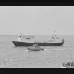 Schwarzweißfoto eines großen Frachtschiffs auf ruhiger See mit einem kleineren Schlepper voraus; im Vordergrund rechts ist ein Schiffsrumpf sichtbar und unten links das Logo des Internationalen Maritimen Museums Hamburg.