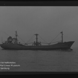 Schwarzweiß-Seitenansicht eines Frachtschiffs mit achterlichem Aufbauten, zwei Ladekranen und leichtem Bugschlag auf ruhiger See; Wasserzeichen des Internationalen Maritimen Museums Hamburg unten links.