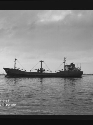 Seitliche Aufnahme eines einsamen Frachtschiffs mit drei Ladebäumen auf ruhiger See unter bewölktem Himmel; im unteren linken Bildrand ein Wasserzeichen des Internationalen Maritimen Museums Hamburg und sichtbarer Filmrand.