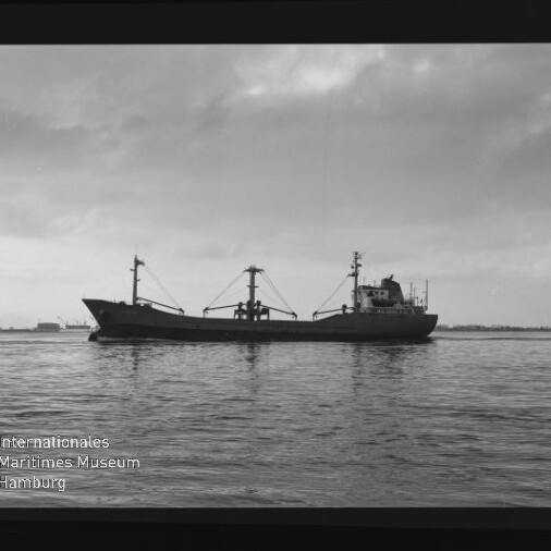 Seitliche Aufnahme eines einsamen Frachtschiffs mit drei Ladebäumen auf ruhiger See unter bewölktem Himmel; im unteren linken Bildrand ein Wasserzeichen des Internationalen Maritimen Museums Hamburg und sichtbarer Filmrand.