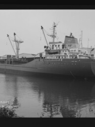 Schwarzweiß-Foto eines beladenen Frachtschiffs am Kai mit mehreren Ladekränen und Lagerhallen im Hintergrund, dessen Rumpf sich ruhig im Hafenwasser spiegelt und mit dem Wasserzeichen des Internationalen Maritimen Museums Hamburg in der unteren linken Ecke.