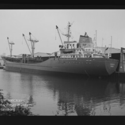 Schwarzweiß-Foto eines beladenen Frachtschiffs am Kai mit mehreren Ladekränen und Lagerhallen im Hintergrund, dessen Rumpf sich ruhig im Hafenwasser spiegelt und mit dem Wasserzeichen des Internationalen Maritimen Museums Hamburg in der unteren linken Ecke.