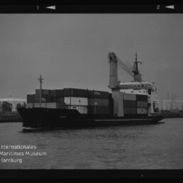 Schwarz-weiß-Aufnahme eines Containerschiffs im Hafen mit gestapelten Frachtcontainern und Bordkran, ruhige Wasserfläche im Vordergrund, sichtbarer Filmrand und Logo des Internationalen Maritimen Museums Hamburg links unten.