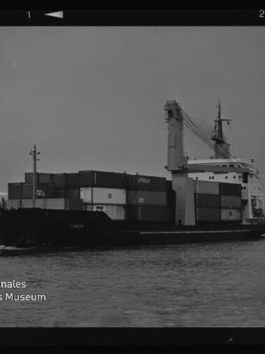 Schwarz-weiß-Aufnahme eines Containerschiffs im Hafen mit gestapelten Frachtcontainern und Bordkran, ruhige Wasserfläche im Vordergrund, sichtbarer Filmrand und Logo des Internationalen Maritimen Museums Hamburg links unten.