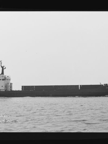 Schwarzweißaufnahme eines langen Frachtschiffs im Seitenprofil auf ruhiger See mit niedrigem Laderaum und Aufbauten am Heck; am Bildrand sichtbare Filmkanten mit Aufdruck AGFA APX 100 und Wasserzeichen des Internationalen Maritimen Museums Hamburg