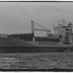 Schwarz-weiß-Seitenansicht des Frachtschiffs MAERSK SEALAND mit Ladekran und Aufbauten am Kai, Vordergrund Wasseroberfläche und links unten das Logo des Internationalen Maritimen Museums Hamburg.
