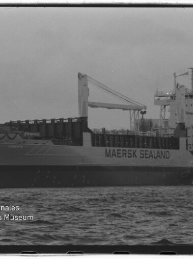 Schwarz-weiß-Seitenansicht des Frachtschiffs MAERSK SEALAND mit Ladekran und Aufbauten am Kai, Vordergrund Wasseroberfläche und links unten das Logo des Internationalen Maritimen Museums Hamburg.