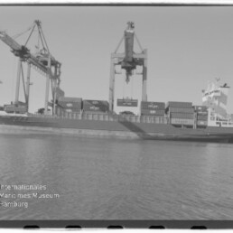 Schwarzweiß-Foto eines am Kai liegenden Containerschiffs mit gestapelten Containern, mehreren großen Hafenkränen darüber und ruhigem Wasser im Vordergrund; unten links das Logo des Internationalen Maritimen Museums Hamburg sichtbar.