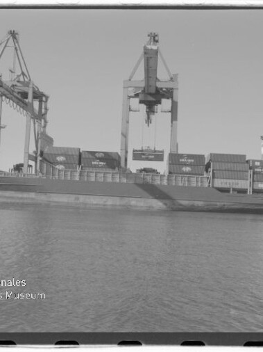 Schwarzweiß-Foto eines am Kai liegenden Containerschiffs mit gestapelten Containern, mehreren großen Hafenkränen darüber und ruhigem Wasser im Vordergrund; unten links das Logo des Internationalen Maritimen Museums Hamburg sichtbar.