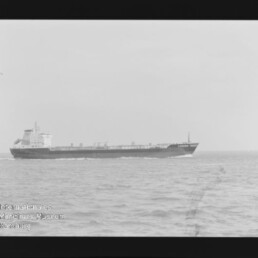 Schwarzweiß-Foto eines langen Frachtschiffs/Tankers auf ruhiger See nahe der Bildmitte, mit sichtbarem Filmrand, der Aufschrift ‚AGFA APX 100‘ am rechten Rand und einem Wasserzeichen des Internationalen Maritimen Museums Hamburg unten links.