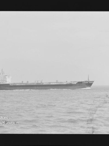 Schwarzweiß-Foto eines langen Frachtschiffs/Tankers auf ruhiger See nahe der Bildmitte, mit sichtbarem Filmrand, der Aufschrift ‚AGFA APX 100‘ am rechten Rand und einem Wasserzeichen des Internationalen Maritimen Museums Hamburg unten links.