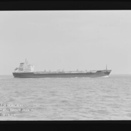 Schwarz-weiß-Foto eines Frachters, der von links nach rechts über eine ruhige See fährt; sichtbare Aufbauten am Heck, hoher Horizont, schwarzer Filmrand mit Markierung und Museumswasserzeichen unten links.