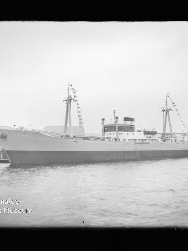 Schwarzweiß-Foto eines fest vertäuten Frachtschiffs mit beflaggten Masten am Kai, ruhiger Wasserfläche und sichtbarem Wasserzeichen des Internationalen Maritimen Museums Hamburg in der linken unteren Ecke.