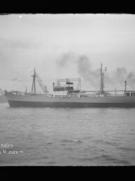 Schwarzweißfoto eines Frachtschiffs im Hafen, seitlich aufgenommen, mit rauchenden Schornsteinen, Masten und Fähnchen; links im Hintergrund eine hohe Hafenbrücke, rechts ein Schlepper, im Vordergrund Wasser und unten links Museumswasserzeichen.