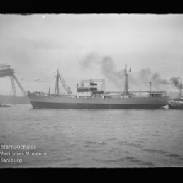 Schwarzweißfoto eines Frachtschiffs im Hafen, seitlich aufgenommen, mit rauchenden Schornsteinen, Masten und Fähnchen; links im Hintergrund eine hohe Hafenbrücke, rechts ein Schlepper, im Vordergrund Wasser und unten links Museumswasserzeichen.