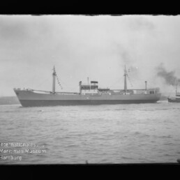 Schwarz-weiß-Foto eines Frachtschiffs mit zwei Masten und Signalflaggen, begleitet von einem rauchenden Schlepper am Heck, auf offener See; unten links ist das Logo des Internationalen Maritimen Museums Hamburg zu sehen.