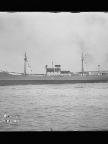Schwarz-weiß-Foto eines Frachtschiffs mit zwei Masten und Signalflaggen, begleitet von einem rauchenden Schlepper am Heck, auf offener See; unten links ist das Logo des Internationalen Maritimen Museums Hamburg zu sehen.