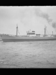 Großes Frachtschiff mit zwei Masten und zentralem Aufbauten, geschmückt mit Festtagsflaggen, fährt im Hafen; rechts begleitet ein Schlepper, der schwarzen Rauch ausstößt, im Vordergrund welliges Wasser und ein bewölkter Himmel.