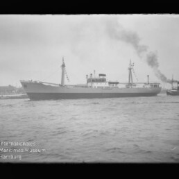 Großes Frachtschiff mit zwei Masten und zentralem Aufbauten, geschmückt mit Festtagsflaggen, fährt im Hafen; rechts begleitet ein Schlepper, der schwarzen Rauch ausstößt, im Vordergrund welliges Wasser und ein bewölkter Himmel.