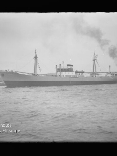 Großes Frachtschiff mit zwei Masten und zentralem Aufbauten, geschmückt mit Festtagsflaggen, fährt im Hafen; rechts begleitet ein Schlepper, der schwarzen Rauch ausstößt, im Vordergrund welliges Wasser und ein bewölkter Himmel.