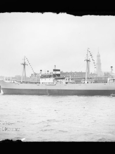 Schwarzweißaufnahme eines großen Schiffs mit zwei Masten und dekorativen Signalflaggen, am Kai liegend; im Vordergrund Wasser und ein kleines Lotsenboot, im Hintergrund eine Stadtsilhouette mit hohem Kirchturm; unten links Wasserzeichen des Internationalen Maritimen Museums Hamburg.