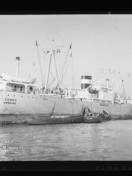 Schwarz‑weiß-Aufnahme eines großen Frachtschiffs mit sichtbar aufgemaltem Namen „Congo Hamburg“, am Kai liegend mit Masten und Ladebäumen, begleitet von Schleppern und Kähnen vor Industriegebäuden.