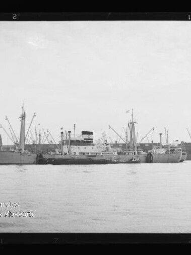 Schwarzweiß-Foto eines Frachtschiffs, das dicht begleitet von einem Schlepper parallel am Hafen liegt, vor einer Kaizeile mit zahlreichen Kränen; unten links sichtbar das Wasserzeichen des Internationalen Maritimen Museums Hamburg.