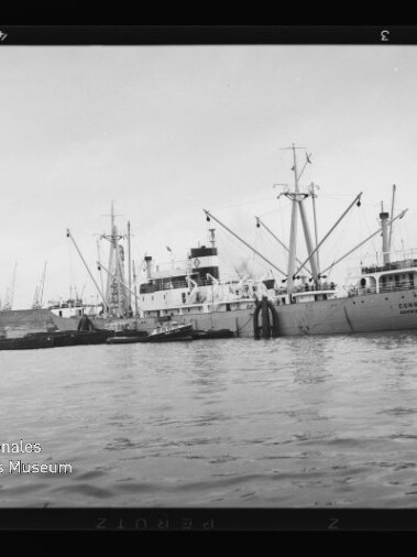 Frachtschiff mit Ladekränen und kleineren Begleitbooten liegt am Kai eines Industriehafens mit einer Reihe von Hafenkränen im Hintergrund; links unten ist das Logo des Internationalen Maritimen Museums Hamburg sichtbar.