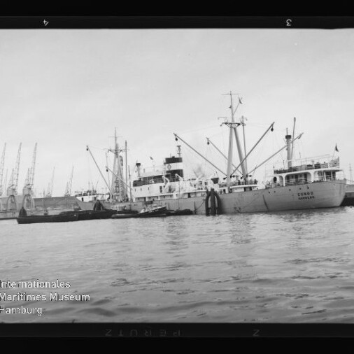 Frachtschiff mit Ladekränen und kleineren Begleitbooten liegt am Kai eines Industriehafens mit einer Reihe von Hafenkränen im Hintergrund; links unten ist das Logo des Internationalen Maritimen Museums Hamburg sichtbar.