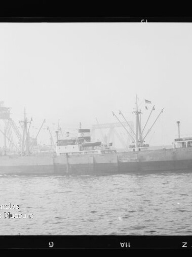 Seitlich abgebildetes Frachtschiff an einem Industriehafen mit mehreren Ladekränen im Hintergrund, ruhige See im Vordergrund und leicht nebliger Dunst.
