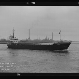 Schwarzweißaufnahme eines Frachtschiffs, das auf ruhigem Wasser vor einer Industrie-Silhouette mit rauchenden Schornsteinen fährt; Filmmarge und Museumswasserzeichen links unten sichtbar.
