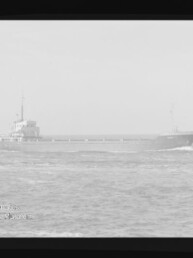 Schwarzweißfoto eines langen Frachtschiffs, seitlich auf ruhiger See fahrend mit sichtbarer Brücke knapp hinter der Bildmitte und Bugwellen, umgeben von einem schwarzen Filmrand links mit der Aufschrift KODAK SAFETY FILM und einem Wasserzeichen des Internationalen Maritimen Museums Hamburg unten links.