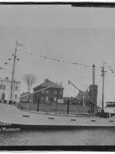 Schwarz-weiß-Foto eines Frachtschiffs mit dem Namen CONRAD am Bug, das mit Wimpelketten geschmückt am Ufer liegt; im Hintergrund Häuser und ein hoher Turm, links unten Wasserzeichen des Internationalen Maritimen Museums Hamburg.