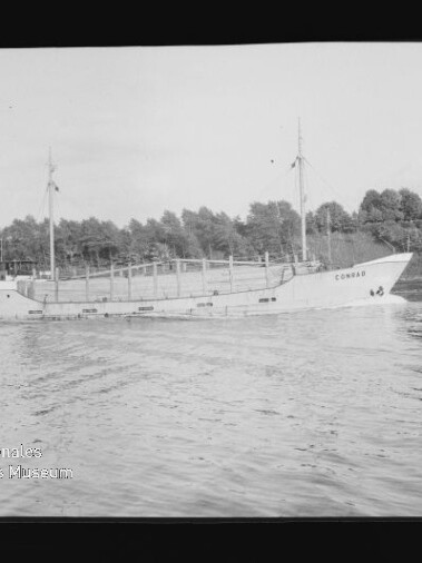 Schwarzweißaufnahme eines Küstenfrachtschiffs mit zwei Masten und dem Schriftzug CONRAD am Bug, das auf ruhigem Wasser vor einer baumbestandenen Uferlinie fährt.