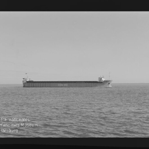 Schwarzweißaufnahme eines langen Frachtschiffs mit lesbarer Rumpfbeschriftung, das ruhig nahe dem Horizont auf ruhiger See fährt; Filmrand und Museumswasserzeichen sind sichtbar.