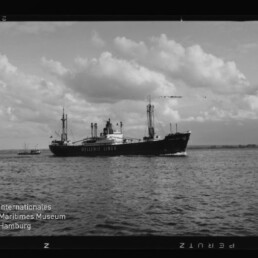 Schwarzweiß-Aufnahme eines Frachtschiffs der Hellenic Lines mit Ladebäumen und Masten, das auf ruhigem Meer unter wolkigem Himmel fährt; Filmbildrand und Museumslogo unten links sichtbar.