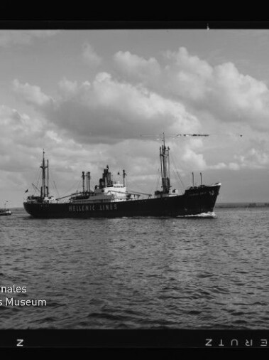 Schwarzweiß-Aufnahme eines Frachtschiffs der Hellenic Lines mit Ladebäumen und Masten, das auf ruhigem Meer unter wolkigem Himmel fährt; Filmbildrand und Museumslogo unten links sichtbar.