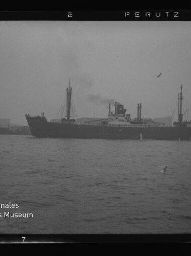 Schwarzweißaufnahme eines großen Dampffrachtschiffs als dunkle Silhouette mittig im Hafen, vor nebliger Hafenanlage mit Schleppern und Möwen, Wasserzeichen des Internationalen Maritimen Museums Hamburg unten links.