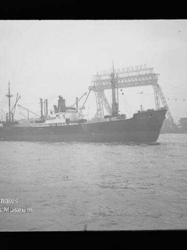 Schwarzweißfotografie eines Frachtschiffs, das in Hafennähe über leicht bewegte See fährt, vor großen Werftkranen und einer hohen Portalkrankonstruktion mit Girlanden, links unten das Logo des Internationalen Maritimen Museums Hamburg.
